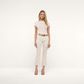 A woman stands against a plain white background, wearing a fitted short-sleeve cream top, the Kendra Cropped Flare Jean in vintage-white, styled with a white belt featuring a gold buckle, and tan high-heeled shoes.