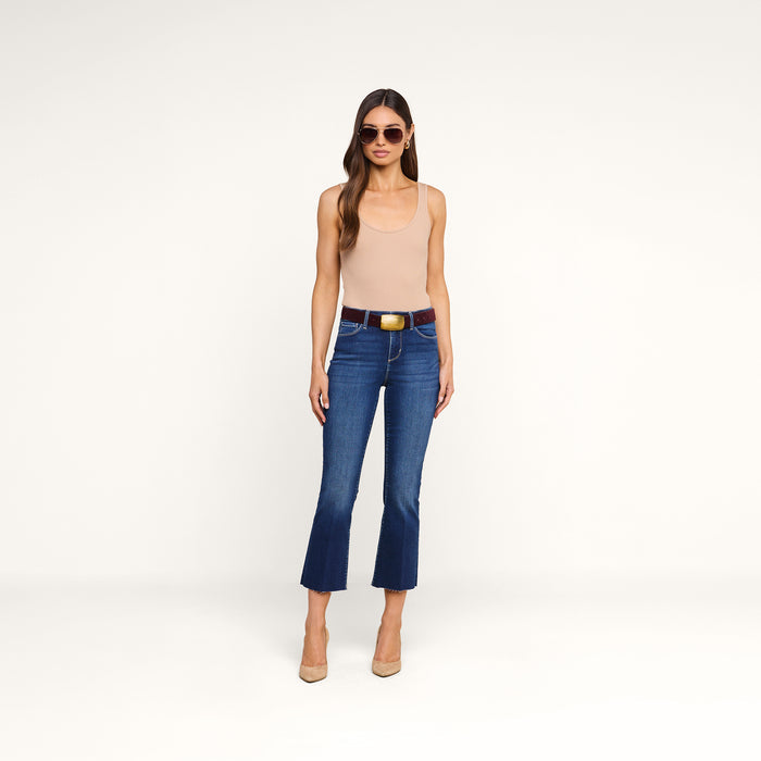 A woman wearing sunglasses, a beige tank top, the Kendra Cropped Flare Jean in blue super stretch denim, a black belt, and beige heels stands against a plain white background.