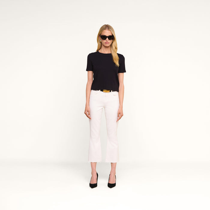 A woman wearing black sunglasses, a black short-sleeve top, and black heels stands against a plain white background. She completes her look with the Kendra Cropped Flare Jean in white stretch denim, featuring a high-rise fit and raw hem.