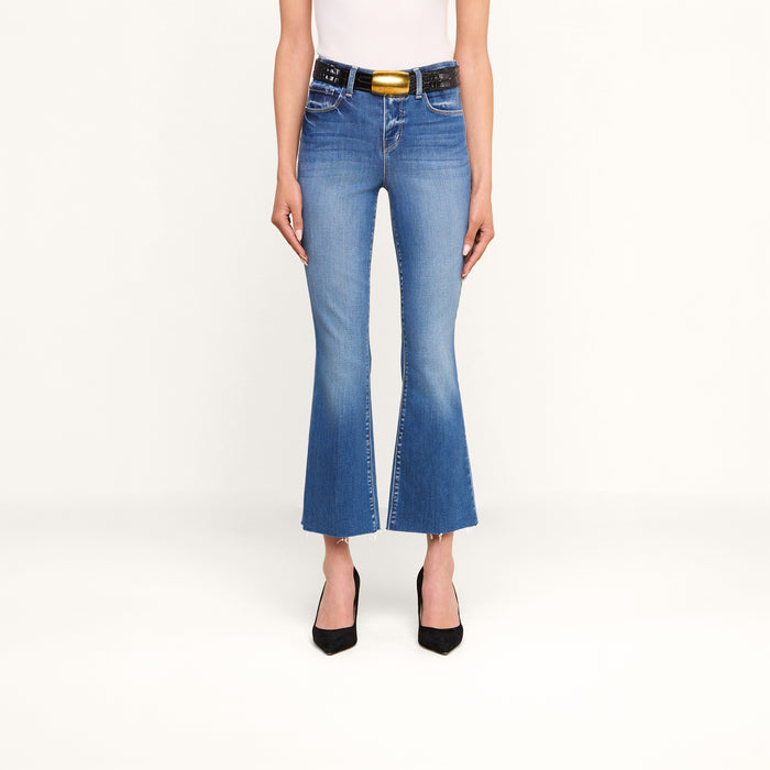 A person wearing the Kendra Cropped Flare Jean—crafted from premium stretch denim—in blue, paired with a black belt featuring a gold buckle, a white top, and black pointed heels, stands against a plain white background. Only the lower half of the body is visible.
