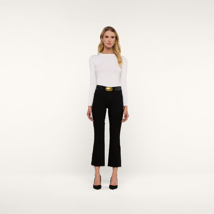 A woman stands against a plain white background, wearing a long-sleeve white top, the Kendra Cropped Flare Jean in black with a belt, and black heels crafted from premium denim.