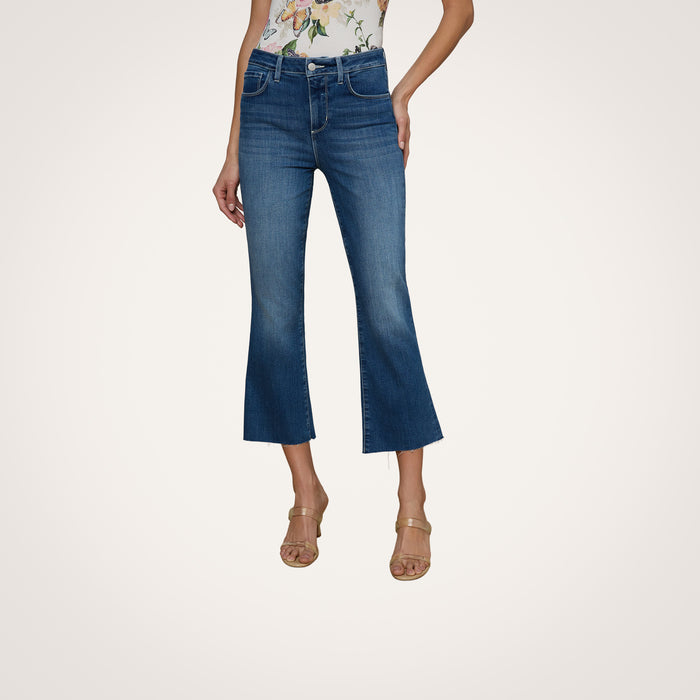 Wearing the Kendra Cropped Flare Jean in a dark denim wash, a person pairs it with a floral sleeveless top and beige heeled sandals while standing against a plain white background. Only the lower half of their body is visible.