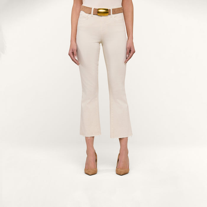 A woman stands facing forward, wearing the Kendra Cropped Flare Jean in vintage white, paired with a tan belt featuring a gold buckle and tan heels. The background is plain white.