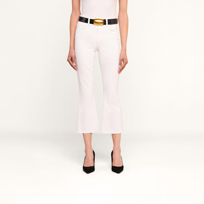 A person wearing the Kendra Cropped Flare Jean in white stretch denim with a raw hem, paired with a white sleeveless top, a black belt featuring a large gold buckle, and black pointed-toe heels stands against a plain white background.