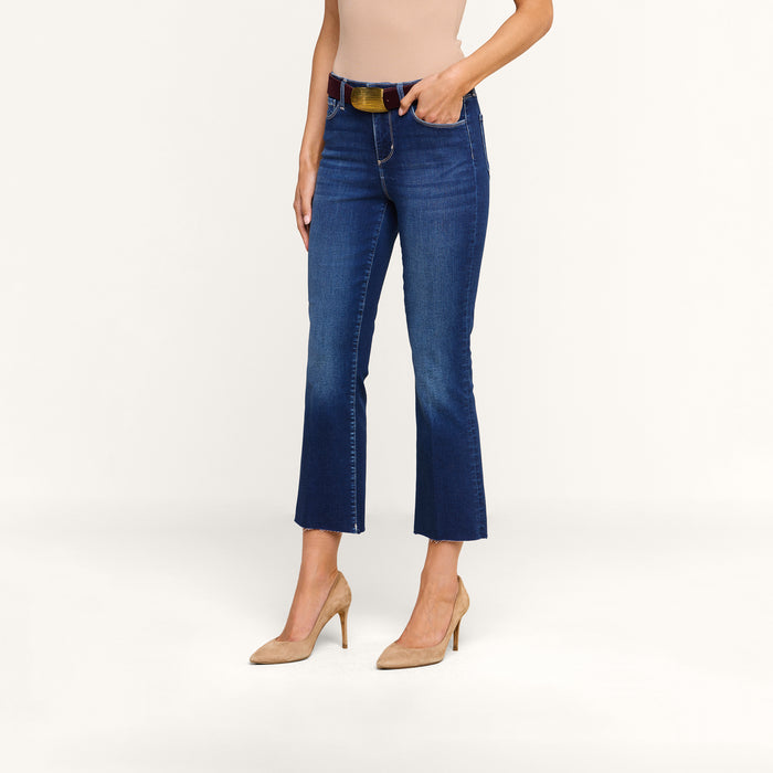 A person wearing the Kendra Cropped Flare Jean in dark blue, styled with a beige top, a black belt, and beige high heels, stands against a plain white background.