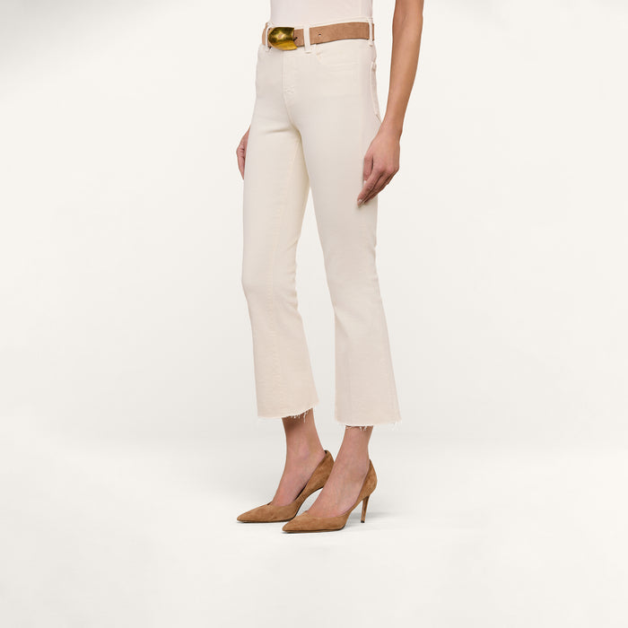 Wearing the Kendra Cropped Flare Jean in vintage white, styled with a tan belt and tan high-heeled shoes, a person stands against a plain white background.