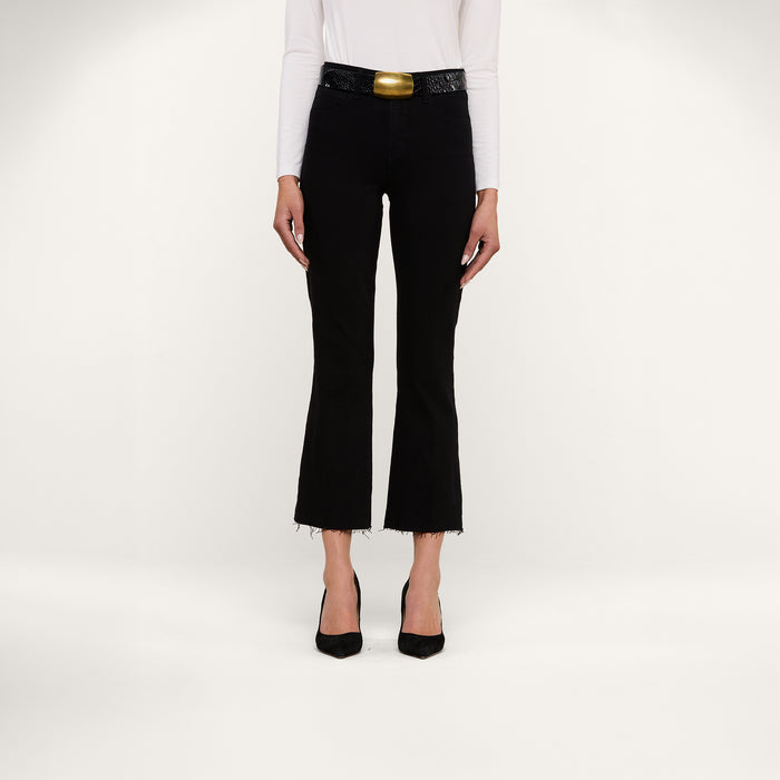 A person wearing a white long-sleeve top, the Kendra Cropped Flare Jean in a rich black wash with frayed hems, a wide black belt featuring a large gold buckle, and black heels stands against a plain white background.