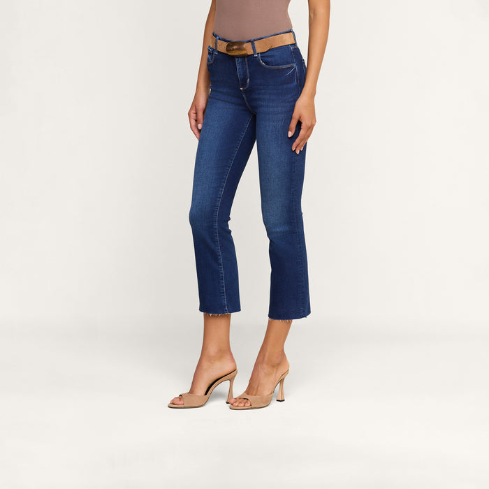 Wearing the Kendra Cropped Flare Jean—crafted from sustainable recycled fiber—a woman pairs her blue cropped flare jeans with a brown sleeveless top, a brown belt, and beige high-heeled shoes as she stands against a plain white background.