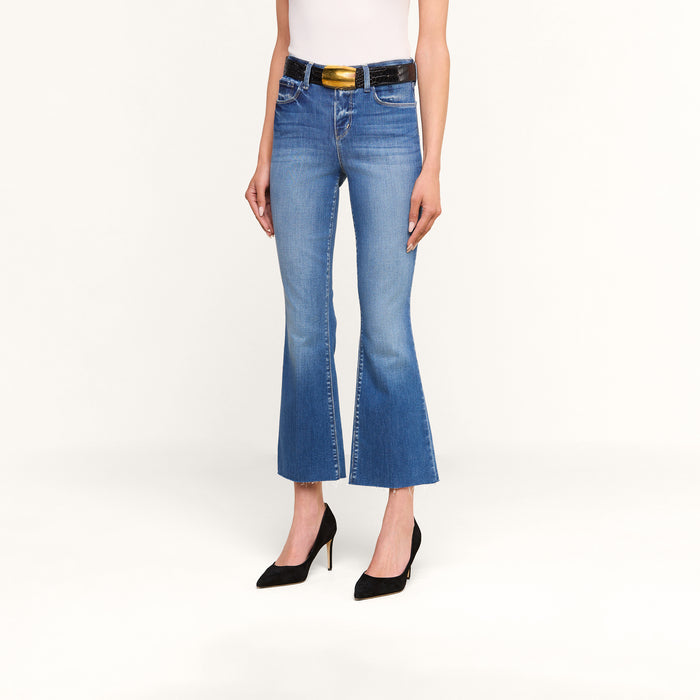 A person wearing the Kendra Cropped Flare Jean in blue premium stretch denim, paired with a black belt featuring a gold buckle, a white top, and black high-heeled shoes, standing against a plain white background.