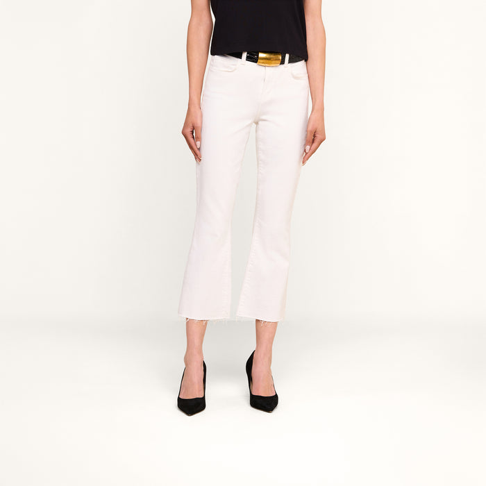 A person wearing the Kendra Cropped Flare Jean in white stretch denim, featuring a high-rise fit, cropped flare leg, and raw hem, paired with a black top and black heels stands against a plain white background. Only the lower half of the body is visible.