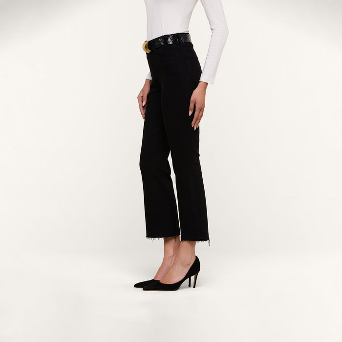 A woman stands in a side profile pose, wearing a white long-sleeve top and the Kendra Cropped Flare Jean—a premium denim, black high-rise style with frayed hems—styled with a black belt and black heeled shoes against a plain light background.