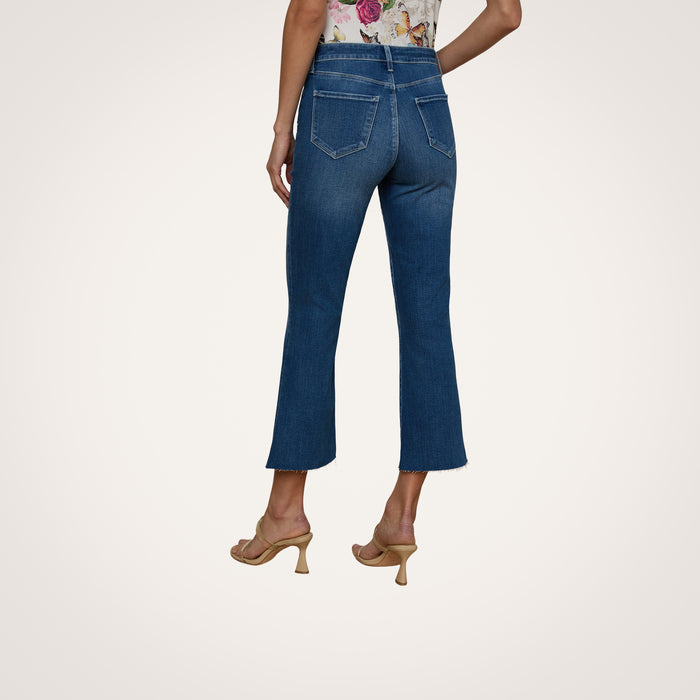 A woman stands facing away against a plain white background, showcasing the back view of the Kendra Cropped Flare Jean in a dark blue denim wash. She pairs the jeans with a floral top and tan heeled sandals.