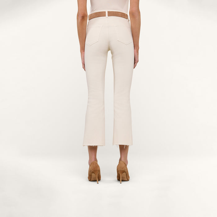 A person stands on a white background, wearing the Kendra Cropped Flare Jean in a vintage-white wash styled with a brown belt and tan high heels. The view is from behind, highlighting the back pockets and frayed hems of the jeans.