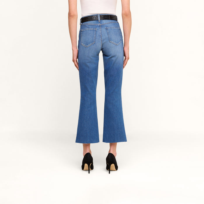 A person stands facing away on a plain white background, wearing the Kendra Cropped Flare Jean in blue—crafted from sustainable recycled fiber—paired with a white sleeveless top, a black belt, and black heeled shoes.