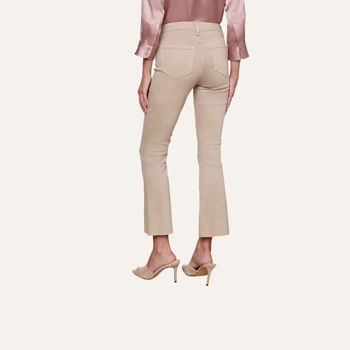 A woman, standing with her back to the camera against a plain background, is wearing Kendra Cropped Flare Jeans in a neutral tan shade, paired with a silky pink blouse and nude high-heeled sandals.