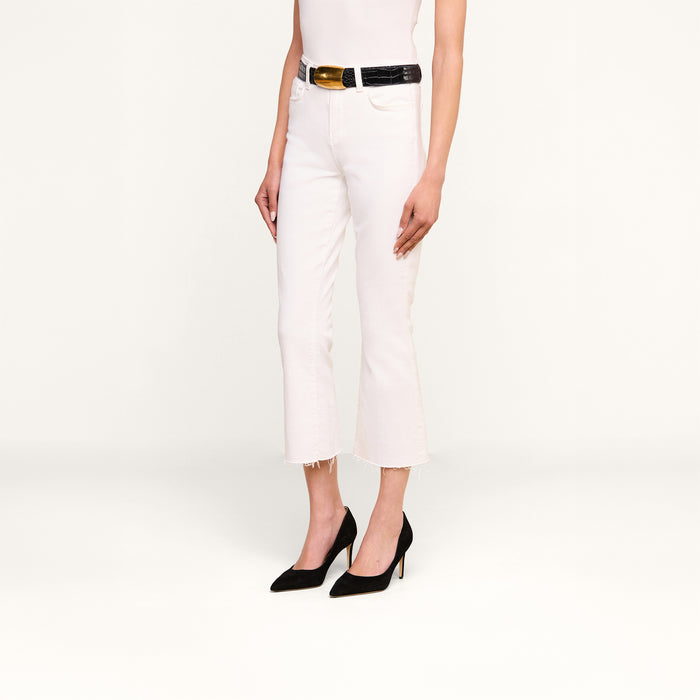 A person wearing the Kendra Cropped Flare Jean in white stretch denim with a raw hem, styled with a black belt, white top, and black pointed-toe heels, stands against a plain white background.
