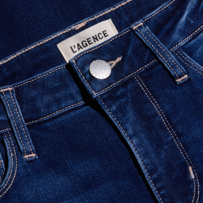 Close-up of the Kendra Cropped Flare Jean in dark blue, featuring a high rise, silver button, front pocket, visible stitching, and a white LAGENCE fabric label above the waistband. Made from super stretch denim for a comfortable fit.