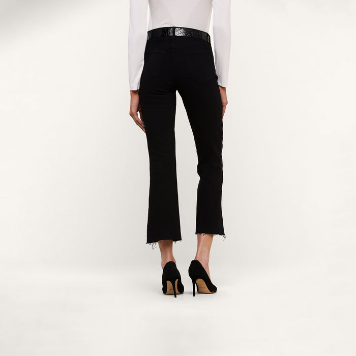 Model shown from the back against a plain light background wearing the Kendra Cropped Flare Jean in black with frayed hems, paired with black high heels and a white long-sleeve top.