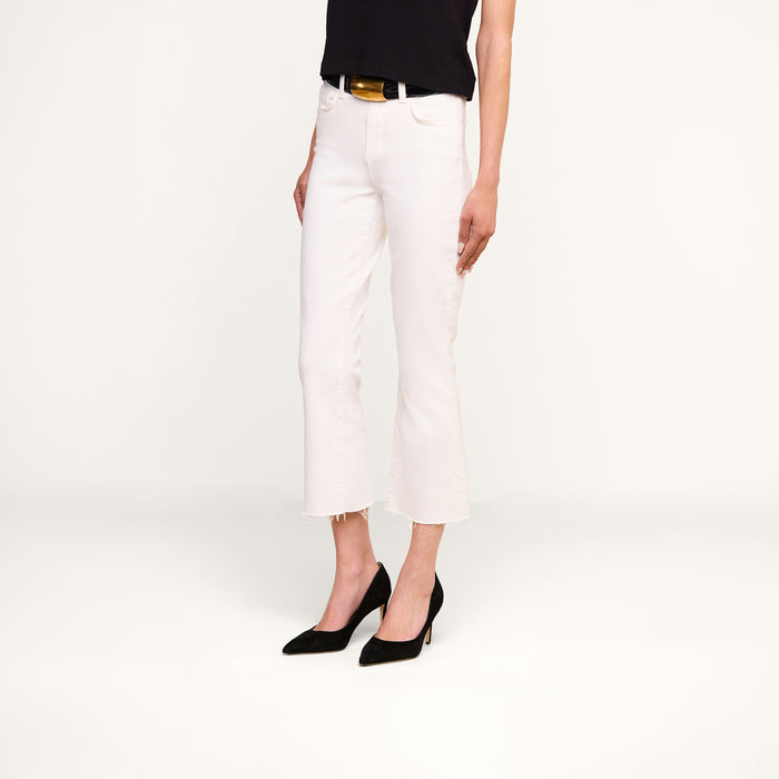 A person wearing the Kendra Cropped Flare Jean in white stretch denim with a raw hem, paired with a black top, a black belt featuring a gold buckle, and black pointed-toe heels stands against a plain white background.
