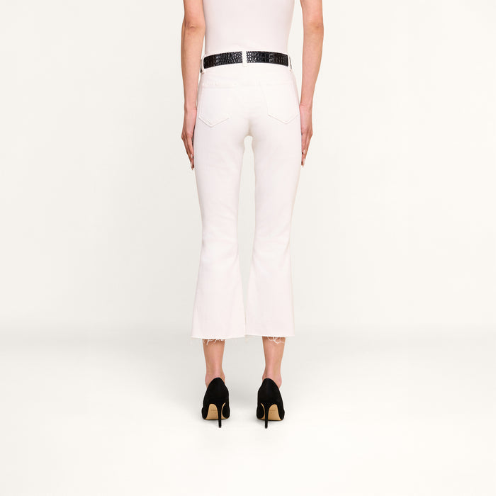 A person stands facing away, wearing the Kendra Cropped Flare Jean in white stretch denim with a raw hem, paired with a black belt and black high-heeled shoes against a plain white background.