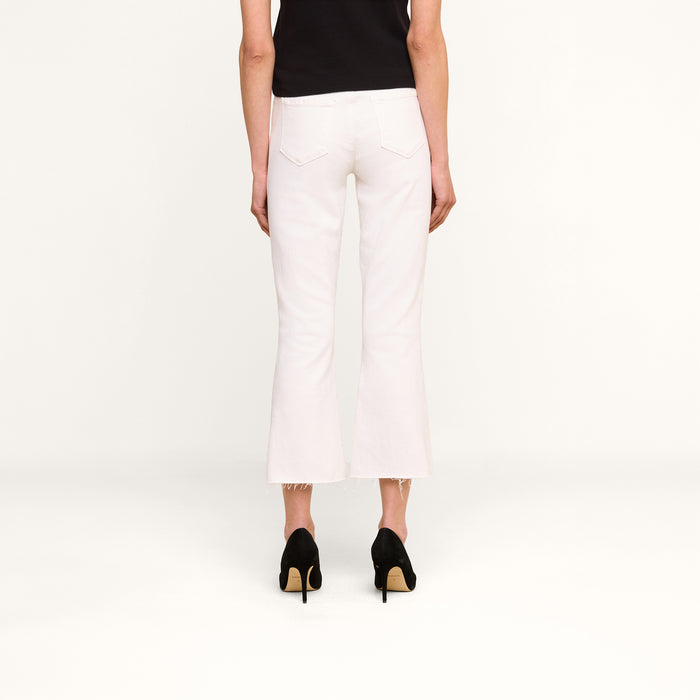 A person is standing facing away, wearing a black top, the Kendra Cropped Flare Jean in white stretch denim featuring a high-rise fit and raw hem, paired with black high-heeled shoes, against a plain white background.