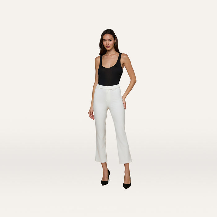 A woman stands against a plain white background, posing with one hand on her hip while wearing a black sleeveless top, black heels, and white Ketra Bootcut Trousers crafted from stretch suit fabric.