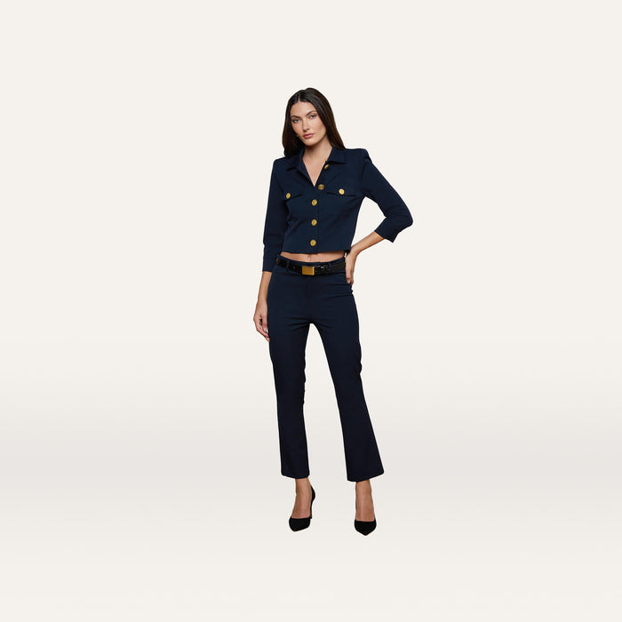 A woman stands against a plain background, wearing the Ketra Bootcut Trouser in midnight navy, paired with a matching tailored jacket featuring gold buttons and crafted from stretch suit fabric. She completes the look with black heels, standing with one hand on her hip and a neutral expression. The baby bootcut silhouette of the Ketra trousers adds a modern touch to her outfit.