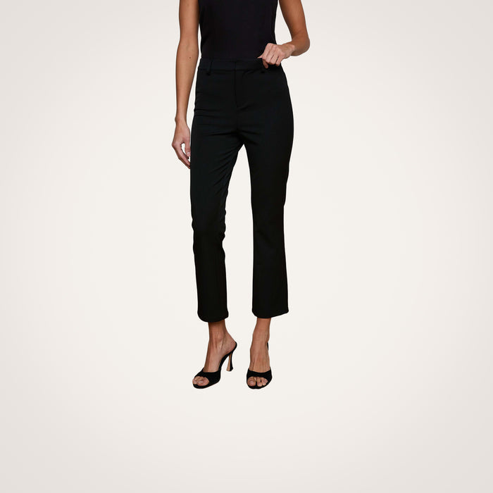 A woman wearing the Ketra Bootcut Trouser in black, featuring an ankle-length baby bootcut silhouette and slant pockets, paired with a sleeveless black top and black high-heeled sandals, stands against a plain light background.