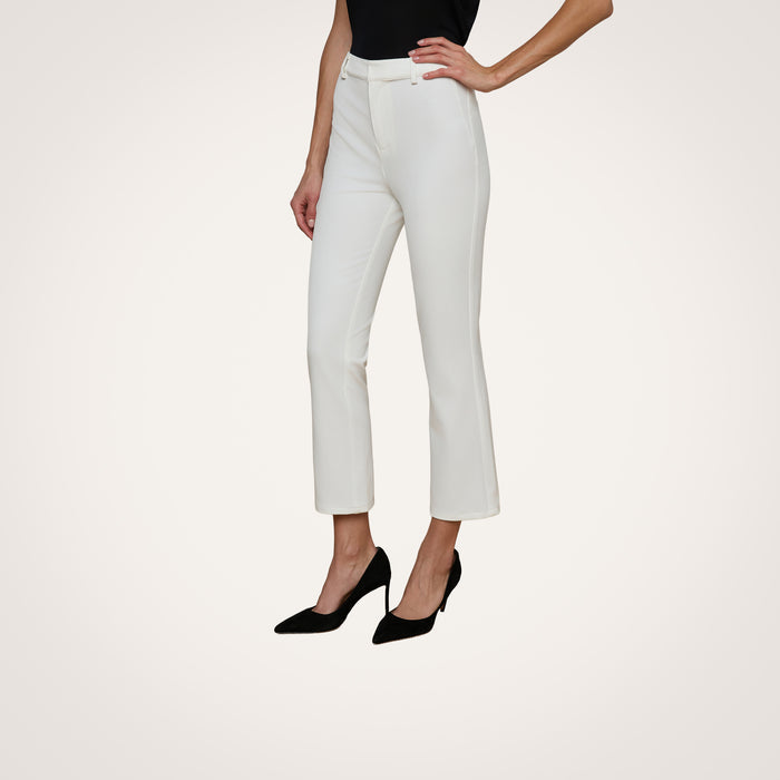 A woman stands with one hand on her hip against a plain white background, wearing the Ketra Bootcut Trouser—white baby bootcut trousers crafted from stretch suit fabric—paired with a black sleeveless top and black high heels.