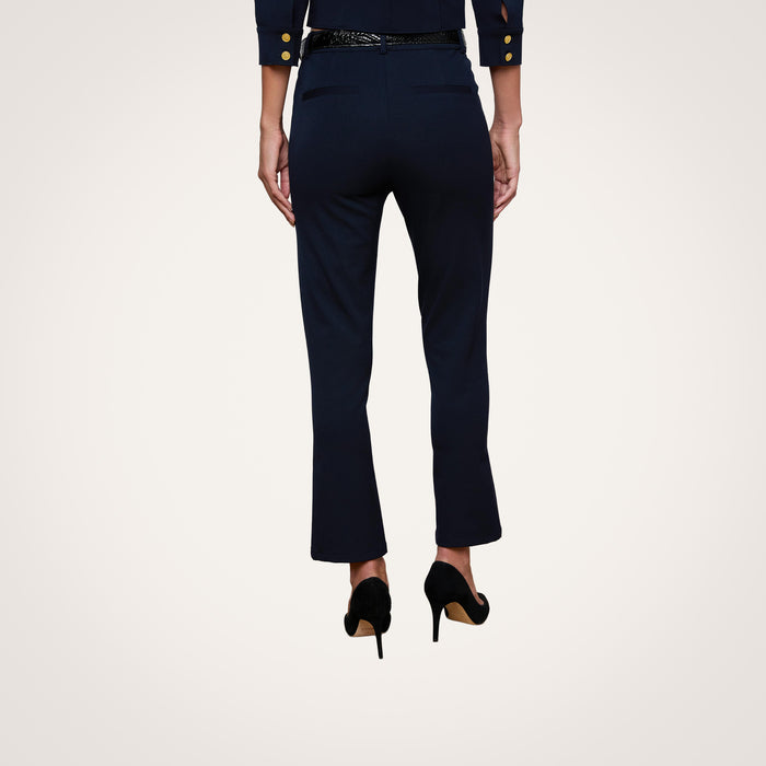 A person stands with their back to the camera, wearing the Ketra Bootcut Trouser in midnight navy and a matching blazer with gold buttons, both crafted from stretch suit fabric, paired with black high heels against a plain white background.