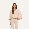 A woman with long brown hair stands facing forward, wearing the beige Kiera Blouse with blouson sleeves and beige high-waisted pants. She has one hand on her hip against a plain white background.