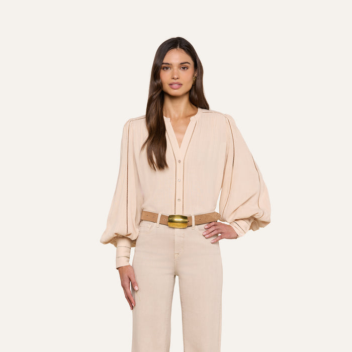 A woman with long brown hair stands facing forward, wearing the beige Kiera Blouse with blouson sleeves and beige high-waisted pants. She has one hand on her hip against a plain white background.