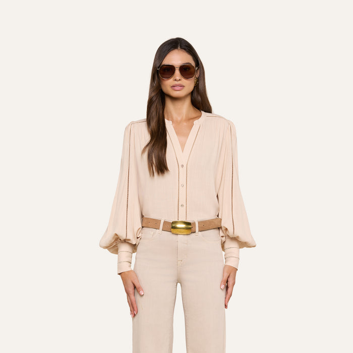 A woman wearing sunglasses, the Kiera Blouse—a beige bohemian top with blouson sleeves—beige pants, and a tan belt stands facing forward against a plain light background.