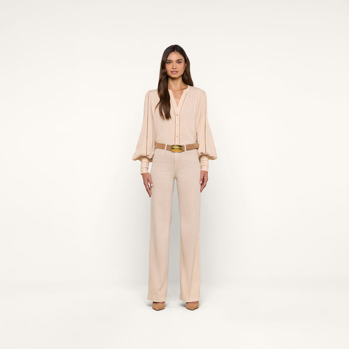 A woman with long, straight brown hair and a neutral expression stands against a plain white background wearing the Kiera Blouse—a beige, bohemian-style top with blouson sleeves—paired with high-waisted beige trousers, a gold belt, and beige heels.