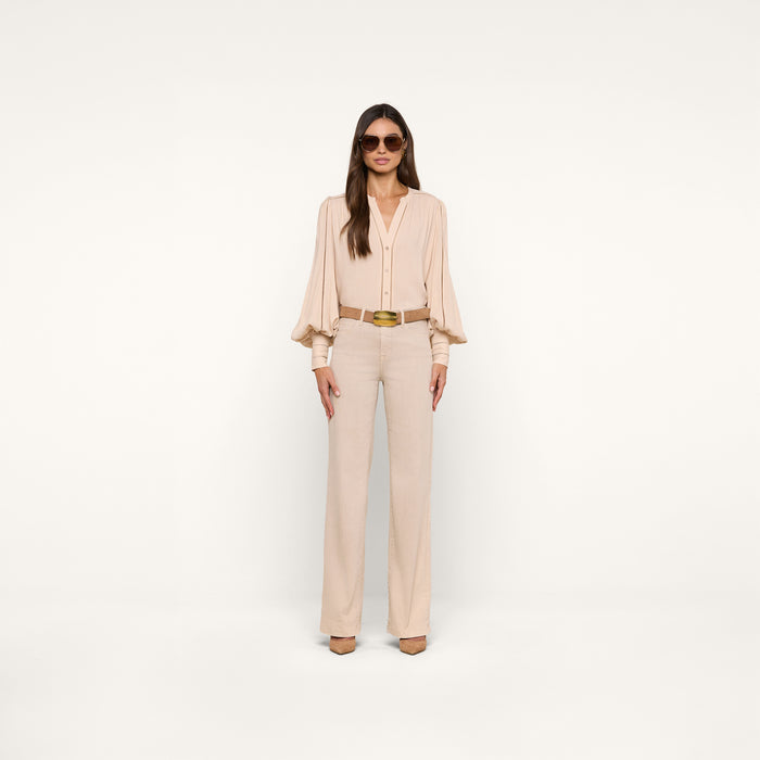 A woman stands confidently in a neutral studio, wearing the Kiera Blouse—a beige bohemian top with blouson sleeves—paired with matching high-waisted trousers, a brown belt, beige heels, and large sunglasses. Her long straight hair and poised stance complete the look.