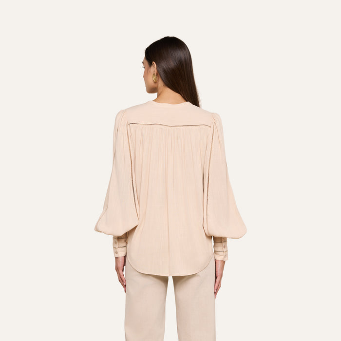A woman stands with her back to the camera, wearing the Kiera Blouse—a loose, light beige bohemian top crafted from textured rayon with long blouson sleeves—paired with matching pants. The background is a plain off-white.