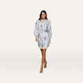 A woman wearing the Kiersten Dress—a light blue and white standout print mini linen dress with blouson sleeves—paired with silver heels, walks forward against a plain white background.