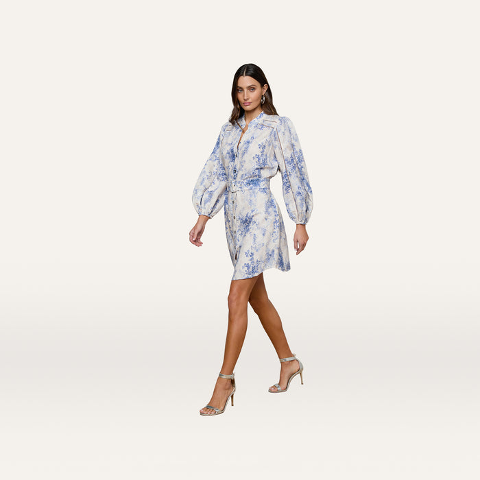 A woman poses in the Kiersten Dress, a light blue and white linen piece with blouson sleeves, a belted waist, and a short hemline. She completes the look with strappy high-heeled sandals against a plain white background.