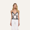 A woman with loose blonde hair and minimal jewelry stands against a plain background, wearing the Kimber Silk Lace Camisole in white with black trim, paired with white pants.