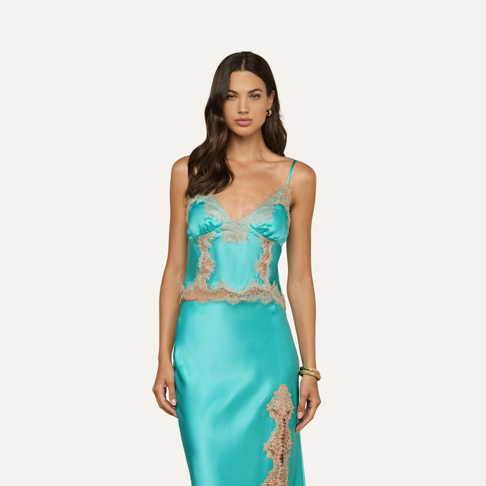 Woman wearing the Kimber Silk Lace Camisole in turquoise, featuring delicate lace trim, standing against a plain light background. She has long, wavy dark hair and accessorizes with gold hoop earrings and a bracelet.