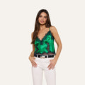 A woman poses against a plain light background with her hands on her hips, modeling the Kimber Silk Lace Camisole in green with black trim and coordinating white pants.