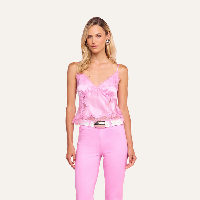 A woman with blonde hair and a neutral expression stands against a pink background wearing the Kimber Silk Lace Camisole, pink pants from the Legally Blonde capsule collection, a white belt, and large round earrings.