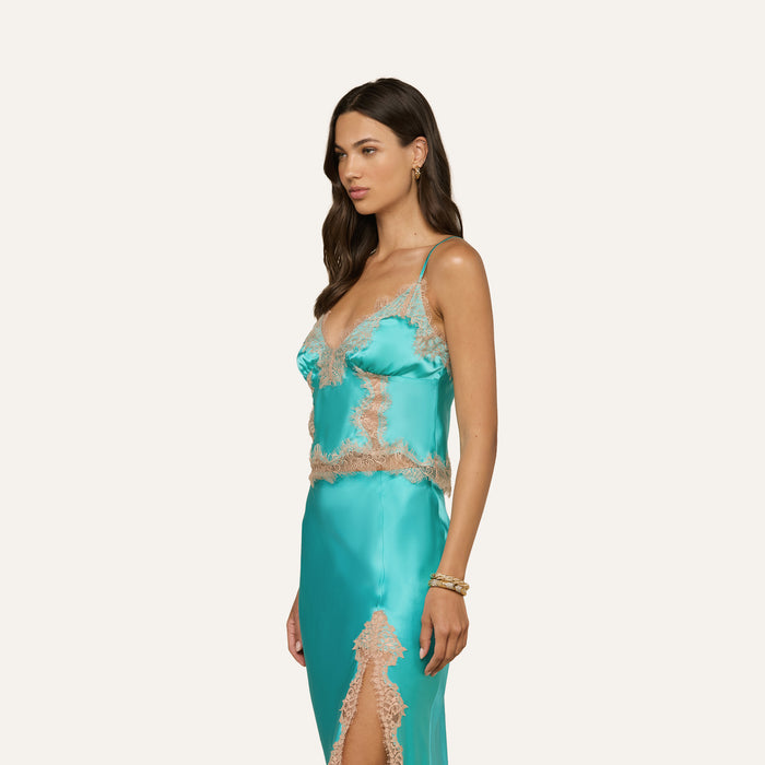 A woman with long brown hair, minimal jewelry, and a calm expression stands sideways against a plain off-white background wearing the Kimber Silk Lace Camisole in turquoise satin with beige lace trim, reminiscent of a delicate slip dress.