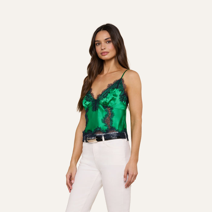 A woman wearing the Kimber Silk Lace Camisole in green, featuring black lace trim, paired with white pants, stands against a plain light background.