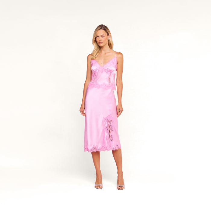 A woman with blonde hair styled to one side poses against a pink backdrop, wearing the Kimber Silk Lace Camisole in light pink from the Legally Blonde capsule collection, paired with thin straps and open-toe heels.