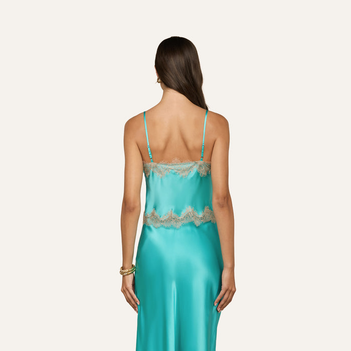 A woman with long brown hair faces away, wearing the Kimber Silk Lace Camisole—a turquoise satin piece featuring thin straps and delicate beige lace trim. She stands against a plain light background.