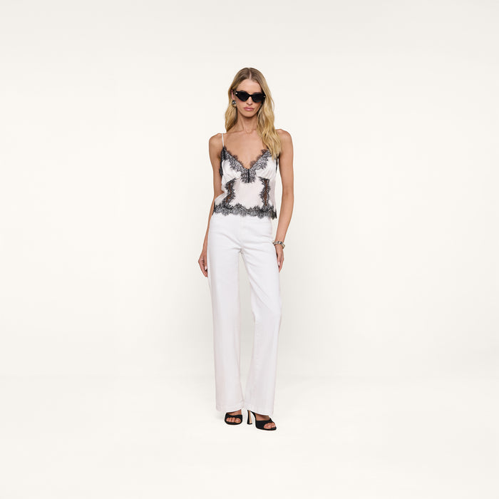 A woman wearing sunglasses, the Kimber Silk Lace Camisole with black detailing, and white pants stands against a plain white background, facing forward with a neutral expression.