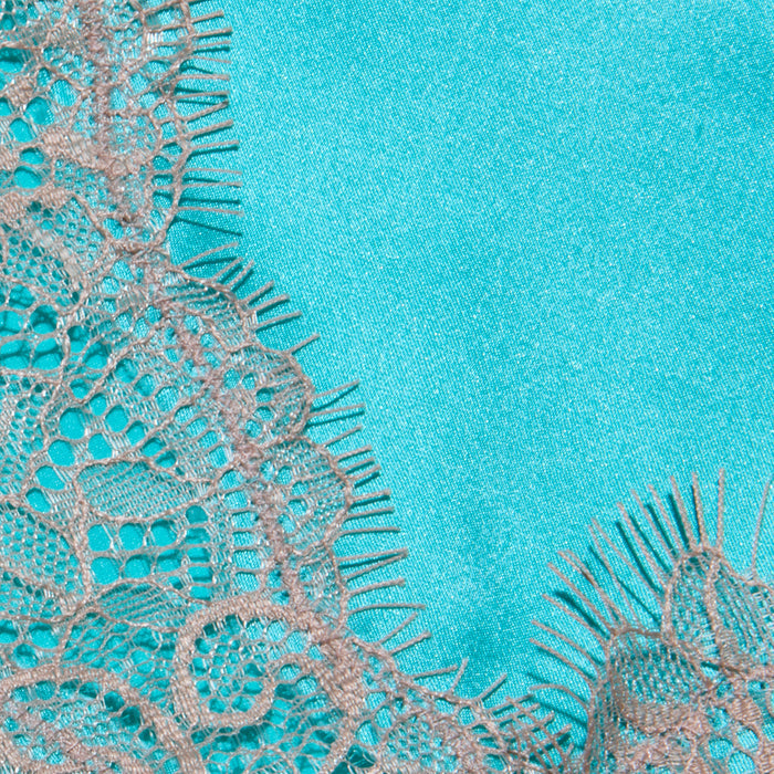 Close-up of the Kimber Silk Lace Camisole, showcasing delicate beige lace with intricate floral patterns against a smooth, bright turquoise background. The camisole’s frayed edges create a soft contrast with the vibrant, solid-colored fabric beneath.