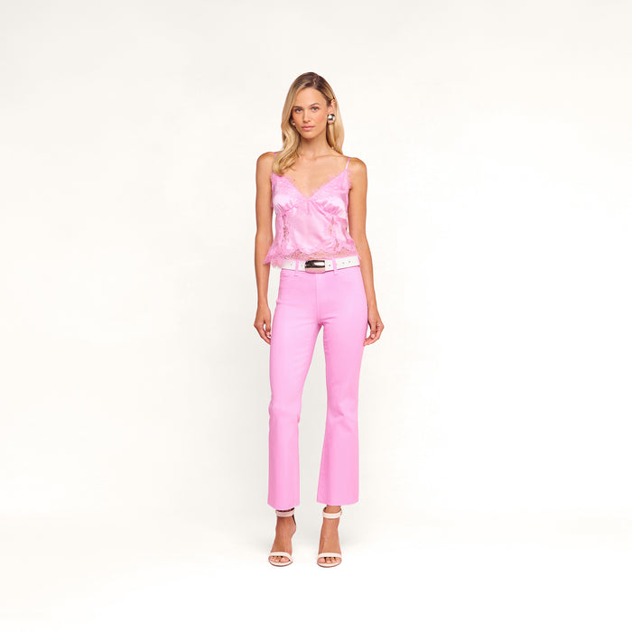 A woman with blonde hair stands against a pink background wearing the Kimber Silk Lace Camisole, paired with light pink pants, a white belt, white heeled sandals, and pearl earrings. She has a neutral expression.