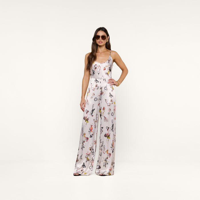 A woman in sunglasses stands confidently against a plain white background, wearing the Kimia Satin Pant—a white, sleeveless jumpsuit featuring a colorful butterfly print and straight-leg pants.